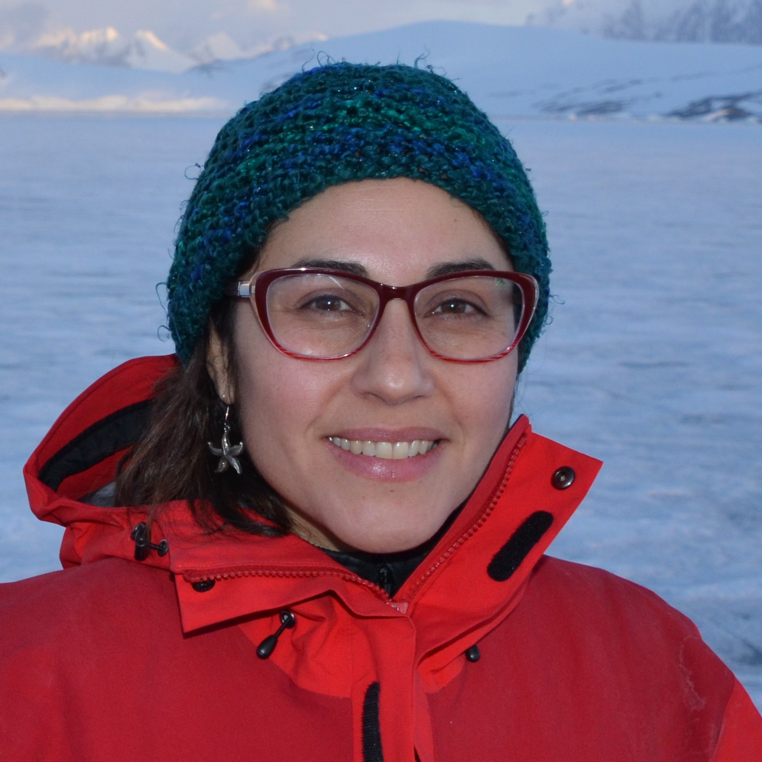 Claudia Maturana Bobadilla in a coat, knit hat, and glasses with a frozen body of water behind her.